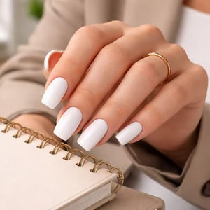 Pode incluir: Uma mão com unhas brancas quadradas e um anel de ouro. A mão segura um caderno bege com encadernação em espiral dourada. A pessoa está usando um blazer bege.