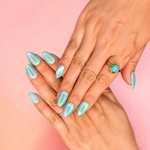 May include: A gold ring featuring a round, polished turquoise stone set in a simple band. The fingernails are shaped into an almond style and are coated in a shimmering, light teal polish with a reflective, cat-eye effect finish.