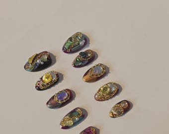 Könnte beinhalten: Eine Auswahl von zehn verzierten Kunstnägeln in Juwelfarben. Jeder Nagel hat ein einzigartiges Design mit goldfarbenen Verzierungen, Perlen und bunten Edelsteinen. Die Nägel sind auf einer weißen Oberfläche angeordnet, um ihre komplizierten Details zu zeigen.
