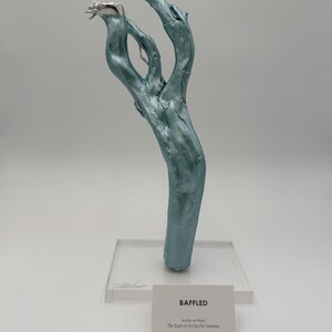 May include: A teal-colored, abstract sculpture made of wood, with silver accents on the tips. The artwork is mounted on a clear acrylic base. A small white card reads "BAFFLED" and "Acrylic on Wood" with the artist's name.