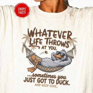 Puede incluir: Una camiseta de manga corta color marfil con un gráfico de un pato con gafas de sol, relajándose en una hamaca entre dos palmeras. El texto dice: WHATEVER LIFE THROWS AT YOU, SOMETIMES YOU JUST GOT TO DUCK. AND KEEP COOL.
