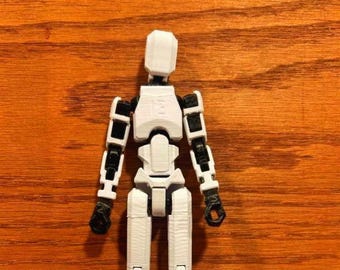 May include: A white articulated action figure with black joints against a wood grain background. The figure has a block-shaped head, segmented limbs, and a detailed torso. The figure is a model or a toy.