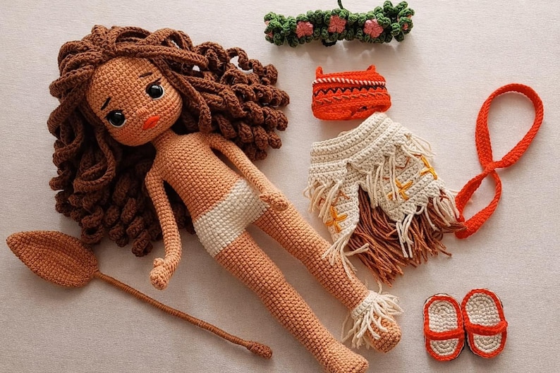 May include: Crocheted doll with brown curly hair, a white diaper, and various accessories. Includes a leaf-shaped paddle, a flower crown, an orange purse, a fringed skirt, a red strap, and red and white sandals. All items are handmade.