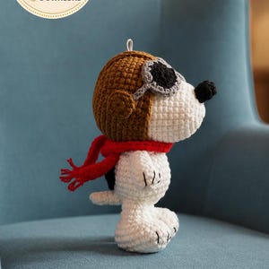 以下が含まれることがあります： 茶色の飛行士ヘルメット、ゴーグル、赤いスカーフを身に着けた、かぎ針編みのスヌーピーの置物。白い体には黒い模様があります。画像には「PDF PATTERN DIGITAL DOWNLOAD」グラフィックが含まれています。