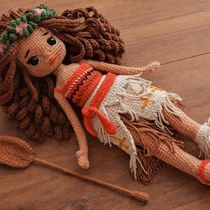 May include: A handmade crochet doll with brown hair, wearing an orange and white outfit, and a floral headband. The doll is lying on a wooden surface, with a crochet oar beside it. The doll's sandals are red and white.