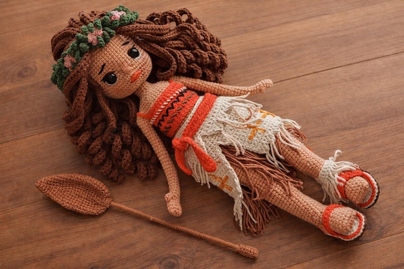 May include: A handmade crochet doll with brown hair, wearing an orange and white outfit, and a floral headband. The doll is lying on a wooden surface, with a crochet oar beside it. The doll's sandals are red and white.