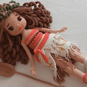 May include: A handmade crochet doll with brown hair, a floral headband, and a red and white outfit. The doll is lying down with a wooden oar beside it. The doll's skin is tan, and it has large, dark eyes.