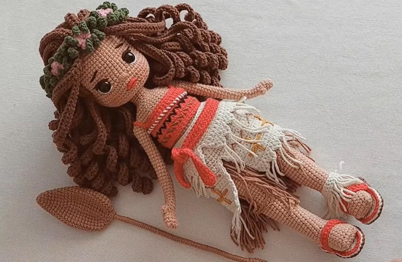 May include: A handmade crochet doll with brown hair, a floral headband, and a red and white outfit. The doll is lying down with a wooden oar beside it. The doll's skin is tan, and it has large, dark eyes.