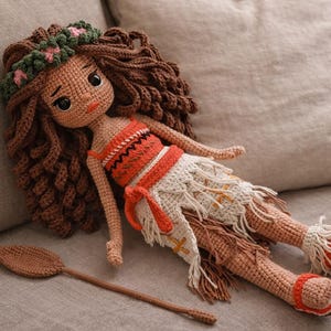 May include: A handmade crochet doll with brown hair, wearing an orange and white outfit, and a floral crown. The doll is lying on a gray surface with a brown crochet oar next to it. The doll's sandals are orange and white.