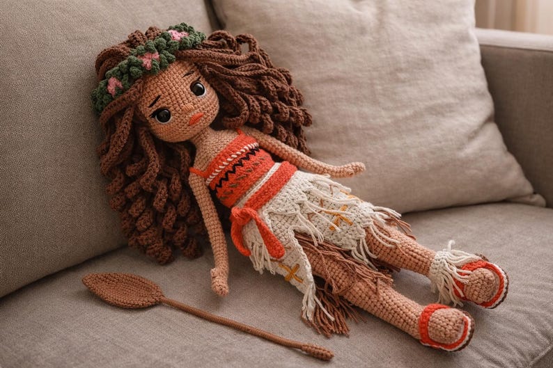 May include: A handmade crochet doll with brown hair, wearing an orange and white outfit, and a floral crown. The doll is lying on a gray surface with a brown crochet oar next to it. The doll's sandals are orange and white.