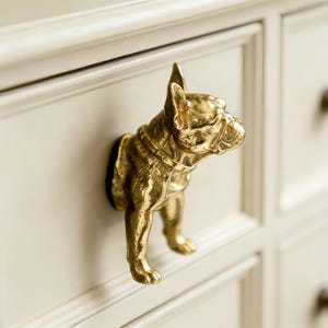 May include: A gold-colored bulldog-shaped drawer pull is mounted on a cream-colored drawer. The bulldog is sculpted with fine details, including a collar. The drawer has a recessed panel design and a dark brown knob.