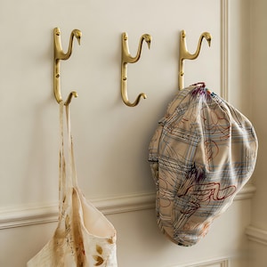 Puede incluir: Tres perchas de pared doradas con forma de cisne, montadas en una pared crema. Una bolsa tote beige con estampado floral cuelga de una percha, y una gorra estampada de otra. Perchas decorativas y funcionales.
