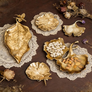 Puede incluir: Una colección de bandejas y platos decorativos dorados con diseños de hojas y botánicos. Una bandeja tiene forma de hoja grande con un adorno de mariposa. Otras bandejas tienen forma de hojas de ginkgo y de loto.