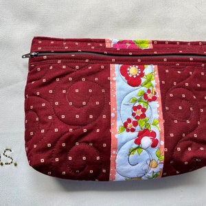 May include: A burgundy quilted zippered pouch with a floral print on one side. The floral print features red, pink, and white flowers with green leaves on a blue background.