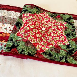 May include: Christmas-themed quilted table runner and star-shaped topper. The runner features stripes with holiday patterns and text, while the topper has a red holly print with a button. The runner is approximately 60 inches long.