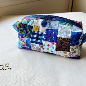 May include: A patchwork zippered pouch with a variety of fabric patterns in shades of blue, green, red, and white. The pouch has a blue zipper and blue ribbon loops on each side. The text "p.s." is embroidered on the white surface.