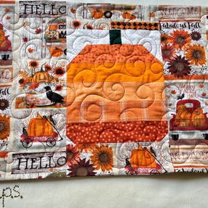 Peut inclure: Chemin de table matelassé sur le thème de l'automne, avec une grande citrouille orange au centre. Le chemin de table présente un motif patchwork avec des tournesols, des citrouilles et les mots "Hello" et "Welcome". La palette de couleurs comprend l'orange, le blanc et le marron.