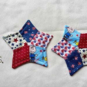 May include: Two star-shaped fabric coasters with a patchwork design. Each coaster features red, white, and blue fabrics with star and patriotic patterns, and a cartoon character. The coasters are sewn together with a button in the centre.