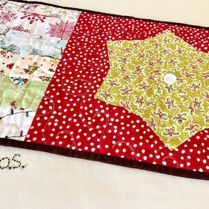 May include: A festive Christmas table runner with a patchwork design. It features a red background with white polka dots, a green star with holly print, and a section with holiday-themed patterns. A white button is in the center of the star.