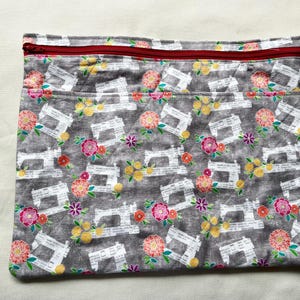 May include: A gray fabric sewing machine case with a red zipper. The case is patterned with white sewing machines and colorful flowers. The case has two pockets and is rectangular in shape.