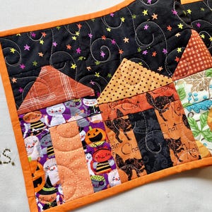 May include: A rectangular quilted table runner featuring a Halloween theme with houses made of fabric patches. The design includes cats, pumpkins, and stars on a black background with orange trim. The letters ps are embroidered in the lower left corner.