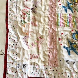 May include: A quilted textile with embroidered designs. The white fabric features cherries, a high-heeled shoe, and blue birds. The border is red with colorful star patterns. The words "DAY" and "TAKE" are visible.
