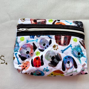 May include: A quilted, zippered pouch with a white background and a colorful dog pattern. The design includes various dog breeds, bones, paw prints, and tennis balls. The pouch has a black zipper and a brown trim.