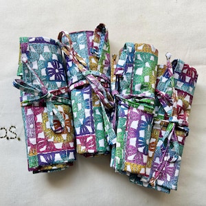 May include: Four fabric rolls featuring a colorful granny square crochet pattern in shades of purple, green, yellow, and blue. Each roll is secured with matching fabric ties. The letters p.s. are embroidered in black thread to the left of the rolls.