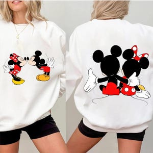 May include: White sweatshirt featuring a Mickey Mouse and Minnie Mouse graphic. The front shows the characters kissing, while the back displays them embracing. Mickey wears red shorts and yellow shoes, Minnie a red polka-dot skirt and bow.