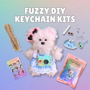 May include: A DIY keychain kit featuring a pink plush bear with sunglasses and a camera. The kit includes accessories, a pipe cleaner, and a small instruction card. The text "FUZZY DIY KEYCHAIN KITS" is at the top.