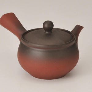 May include: A ceramic teapot with a dark brown lid and a reddish-brown body. The teapot has a side handle and a spout. The lid has a small, round knob. The teapot is designed for brewing tea.