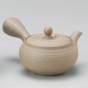 Puede incluir: Una tetera de cerámica beige con un asa lateral y una tapa con un pequeño pomo. La tetera tiene un cuerpo redondeado y un pico para verter. La superficie tiene un aspecto texturizado y ligeramente rugoso.