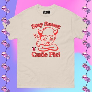 Op de afbeelding: Een crèmekleurig t-shirt met een rode duivelafbeelding en de tekst "Stay Sweet Cutie Pie!". De afbeelding toont een knipogende duivel met hoorns en een staart. Het shirt heeft een ronde hals en korte mouwen.