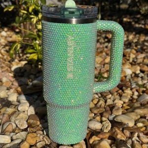 May include: A sparkling green tumbler with a handle, covered in iridescent rhinestones. The tumbler has a clear lid and a light green slider. The handle is also covered in rhinestones. The tumbler is sitting on a bed of rocks.