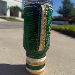 Puede incluir: Un vaso adornado con pedrer&iacute;a verde, dorada y blanca. El vaso est&aacute; cubierto de pedrer&iacute;a verde con rayas doradas y blancas. La parte superior e inferior del vaso tienen detalles dorados y blancos. El vaso es cil&iacute;ndrico y tiene una tapa transparente.