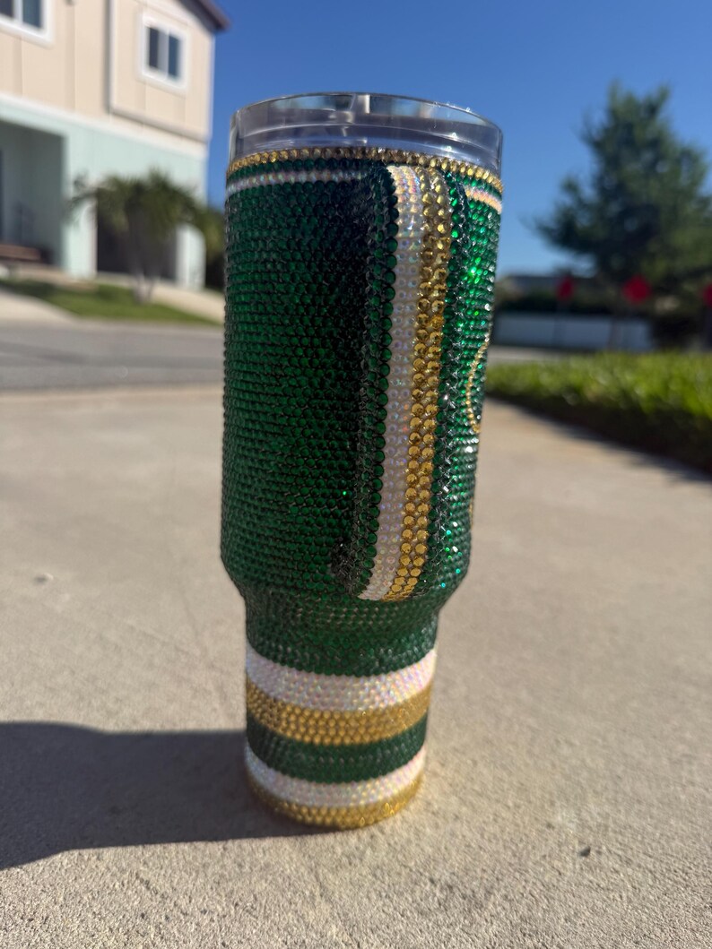 Puede incluir: Un vaso adornado con pedrer&iacute;a verde, dorada y blanca. El vaso est&aacute; cubierto de pedrer&iacute;a verde con rayas doradas y blancas. La parte superior e inferior del vaso tienen detalles dorados y blancos. El vaso es cil&iacute;ndrico y tiene una tapa transparente.