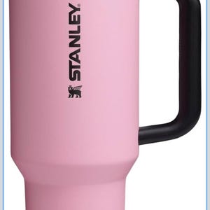 Puede incluir: Un vaso t&eacute;rmico Stanley rosa con asa y tapa negras. El vaso tiene el logotipo de Stanley y las palabras "STAY LATE START EARLY" impresas en la tapa.