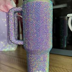 May include: A large, iridescent tumbler covered in small, sparkling rhinestones. The tumbler has a handle and a black lid. The color shifts between purple, blue, and green. The tumbler is sitting on a wooden surface.