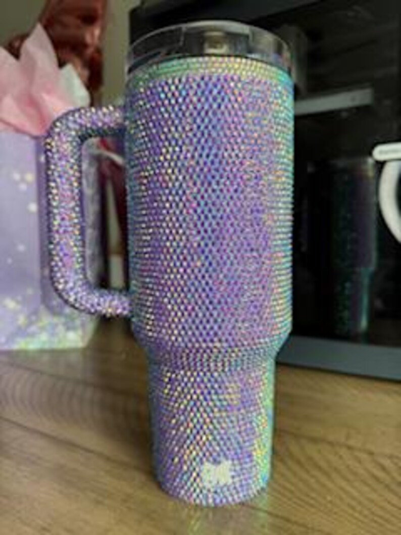 May include: A large, iridescent tumbler covered in small, sparkling rhinestones. The tumbler has a handle and a black lid. The color shifts between purple, blue, and green. The tumbler is sitting on a wooden surface.