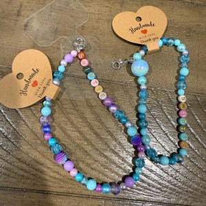 May include: Two beaded lanyards with a variety of colorful beads in shades of blue, purple, and pink. One lanyard has the word "Selfie" spelled out in letter beads. Each lanyard is attached to a heart-shaped tag that reads "Handmade with love, Thank you."