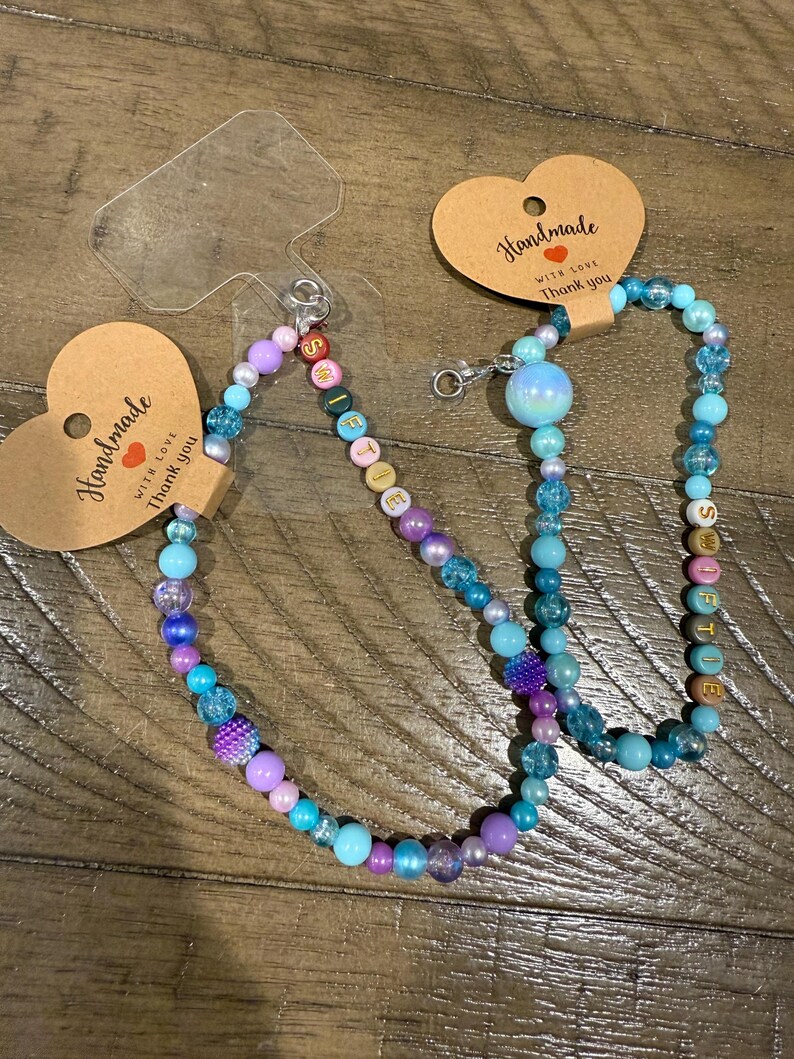 May include: Two beaded lanyards with a variety of colorful beads in shades of blue, purple, and pink. One lanyard has the word "Selfie" spelled out in letter beads. Each lanyard is attached to a heart-shaped tag that reads "Handmade with love, Thank you."