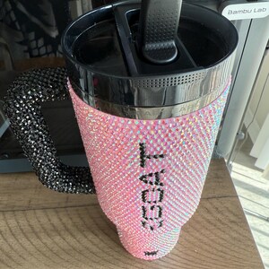 Puede incluir: Un vaso rosa con pedrer&iacute;a, con asa y tapa negras. El vaso est&aacute; cubierto de pedrer&iacute;a iridiscente y presenta el texto "I GOAT" en negro. El asa tambi&eacute;n est&aacute; cubierta de pedrer&iacute;a negra.