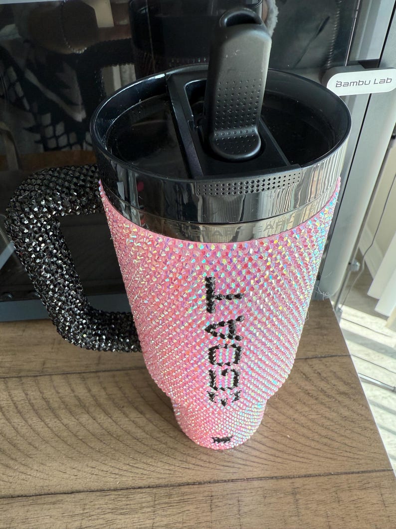 Puede incluir: Un vaso rosa con pedrer&iacute;a, con asa y tapa negras. El vaso est&aacute; cubierto de pedrer&iacute;a iridiscente y presenta el texto "I GOAT" en negro. El asa tambi&eacute;n est&aacute; cubierta de pedrer&iacute;a negra.