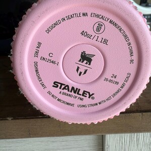 Puede incluir: Primer plano de la base de una botella de agua Stanley rosa. El texto dice "Designed in Seattle WA" y "40oz/1.18L". El logotipo de Stanley est&aacute; en el centro. La botella es libre de BPA y apta para lavavajillas.