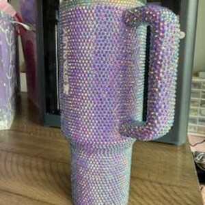 May include: A large, bedazzled tumbler with a handle, covered in iridescent rhinestones. The tumbler is primarily purple, with a clear lid and a handle. The rhinestones catch the light, creating a sparkling effect. The tumbler sits on a wooden surface.