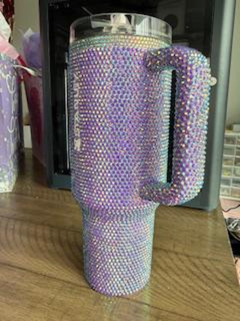 May include: A large, bedazzled tumbler with a handle, covered in iridescent rhinestones. The tumbler is primarily purple, with a clear lid and a handle. The rhinestones catch the light, creating a sparkling effect. The tumbler sits on a wooden surface.