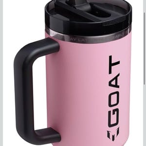 Puede incluir: Vaso aislado rosa con asa y tapa negras. El vaso tiene la palabra "GOAT" impresa verticalmente en negro. Un logotipo negro en la parte inferior. Dise&ntilde;ado para mantener las bebidas fr&iacute;as o calientes durante horas.