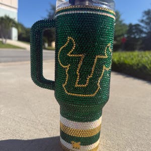 Puede incluir: Un vaso verde y dorado adornado con pedrer&iacute;a con asa. El vaso presenta la letra "U" y una "S" estilizada en dorado. La parte inferior del vaso tiene rayas horizontales blancas, doradas y verdes. El asa tambi&eacute;n est&aacute; cubierta de pedrer&iacute;a verde.