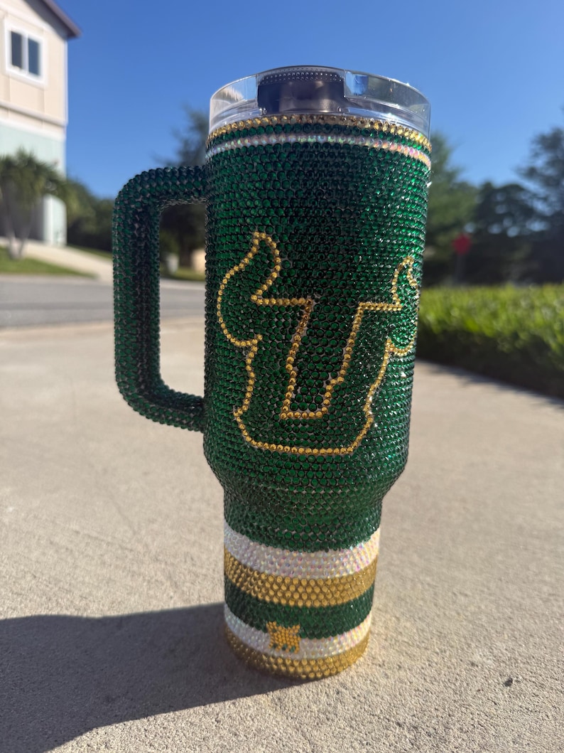 Puede incluir: Un vaso verde y dorado adornado con pedrer&iacute;a con asa. El vaso presenta la letra "U" y una "S" estilizada en dorado. La parte inferior del vaso tiene rayas horizontales blancas, doradas y verdes. El asa tambi&eacute;n est&aacute; cubierta de pedrer&iacute;a verde.