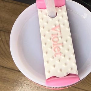 K&ouml;nnte beinhalten: Eine wei&szlig;e und rosa Badebombenform mit dem Wort "Violet" in Rosa. Die Form hat eine strukturierte Oberfl&auml;che mit kleinen, erhabenen Punkten. Die Form ist teilweise in einer wei&szlig;en Sch&uuml;ssel versenkt.